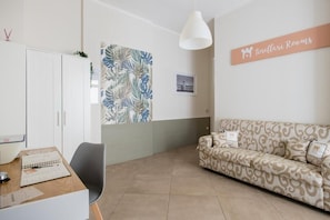 A room with a white wardrobe, a patterned sofa, a desk with a chair, and a wall-mounted sign reading 'Tinellari Rooms'. - Tinellari Rooms (Naples)
