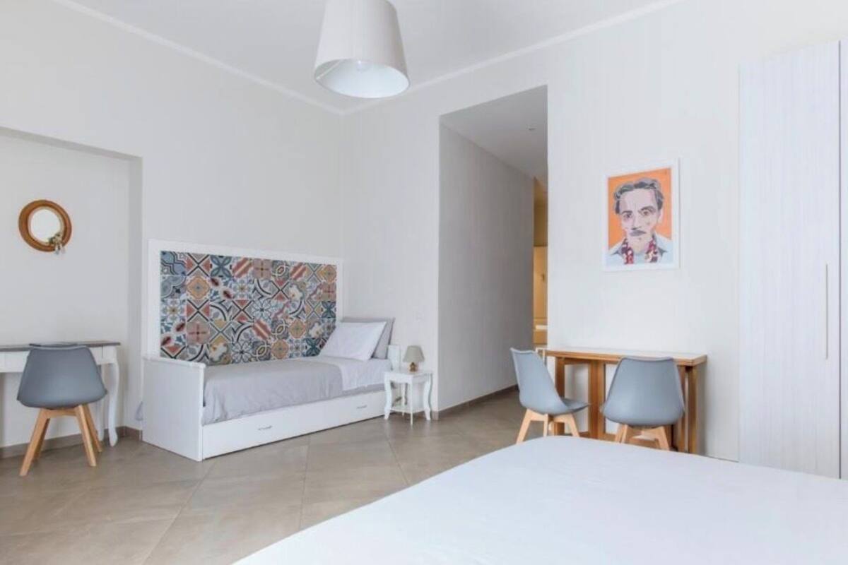 A modern bedroom with a bed, a desk, two chairs, a small table, and a wall-mounted artwork.