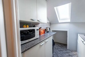 Eine kleine Küche mit weißen Schränken, einer Mikrowelle, einem Toaster und einem Wasserkocher. Ein kleines Fenster sorgt für natürliches Licht.