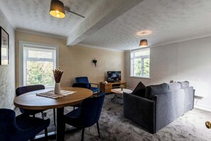 A cozy living room with a round wooden table, blue chairs, a grey sofa, and a television on a wooden stand. - St Andrew's by Guestz (Lytham)