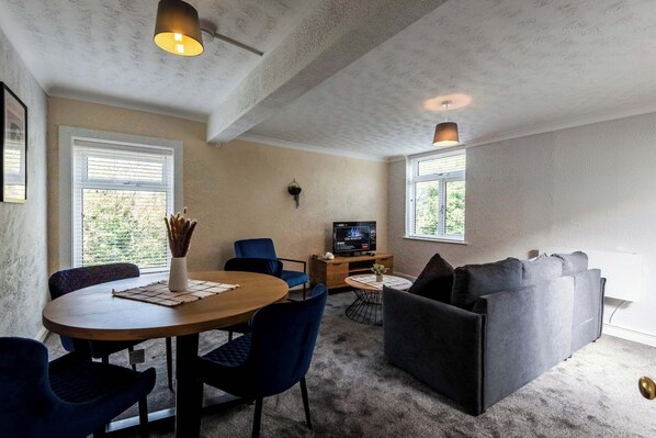 Ein gemütliches Wohnzimmer mit einem runden Holztisch, blauen Stühlen, einem grauen Sofa und einem Fernseher auf einem Holzschrank.