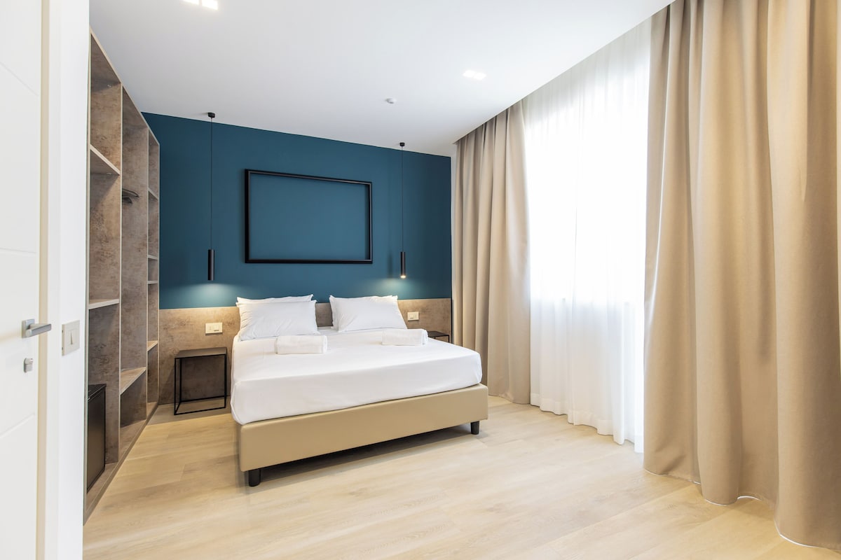 A modern bedroom with a large bed, a nightstand, a wall-mounted light, and a window with curtains.