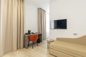 A modern hotel room with a desk, chair, sofa, and a flat-screen TV mounted on the wall.