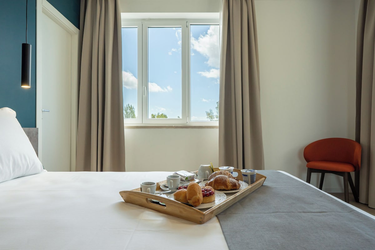 A hotel room with a bed, a breakfast tray with pastries and coffee, a chair, and a window with curtains.