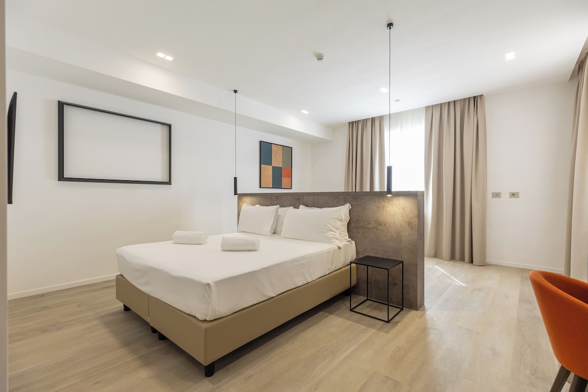 A modern hotel room with a large bed, a bedside table, a chair, and a view of the outdoors.