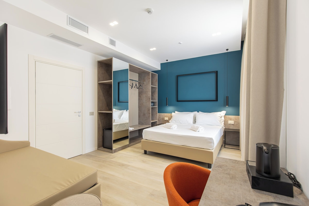 A modern hotel room with a bed, a seating area, and a wardrobe.