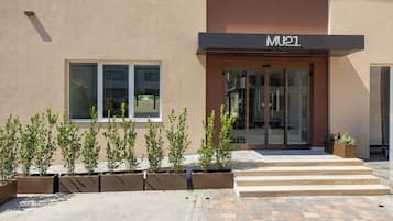Un bâtiment moderne avec une enseigne indiquant « MU1 » au-dessus de l’entrée. Des plantes en pot sont disposées devant l’entrée, et un espace pavé mène à la porte.