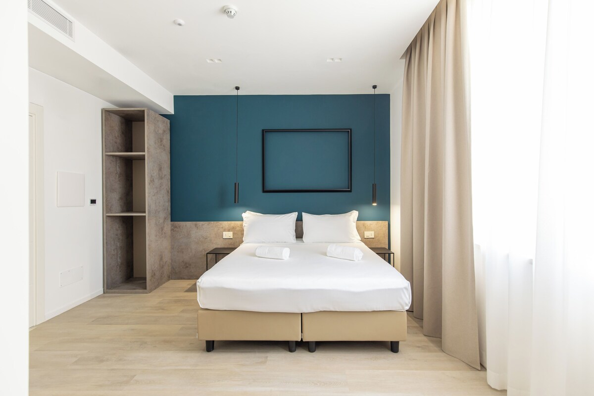 A modern bedroom with a large bed, a wardrobe, a bedside table, and a wall-mounted light fixture.