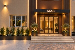 A modern hotel entrance with a nameplate reading 'MU21,' a reception area, and potted plants.