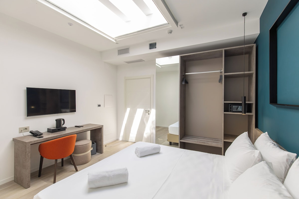 A modern hotel room with a bed, a desk, a chair, a TV, and a wardrobe.