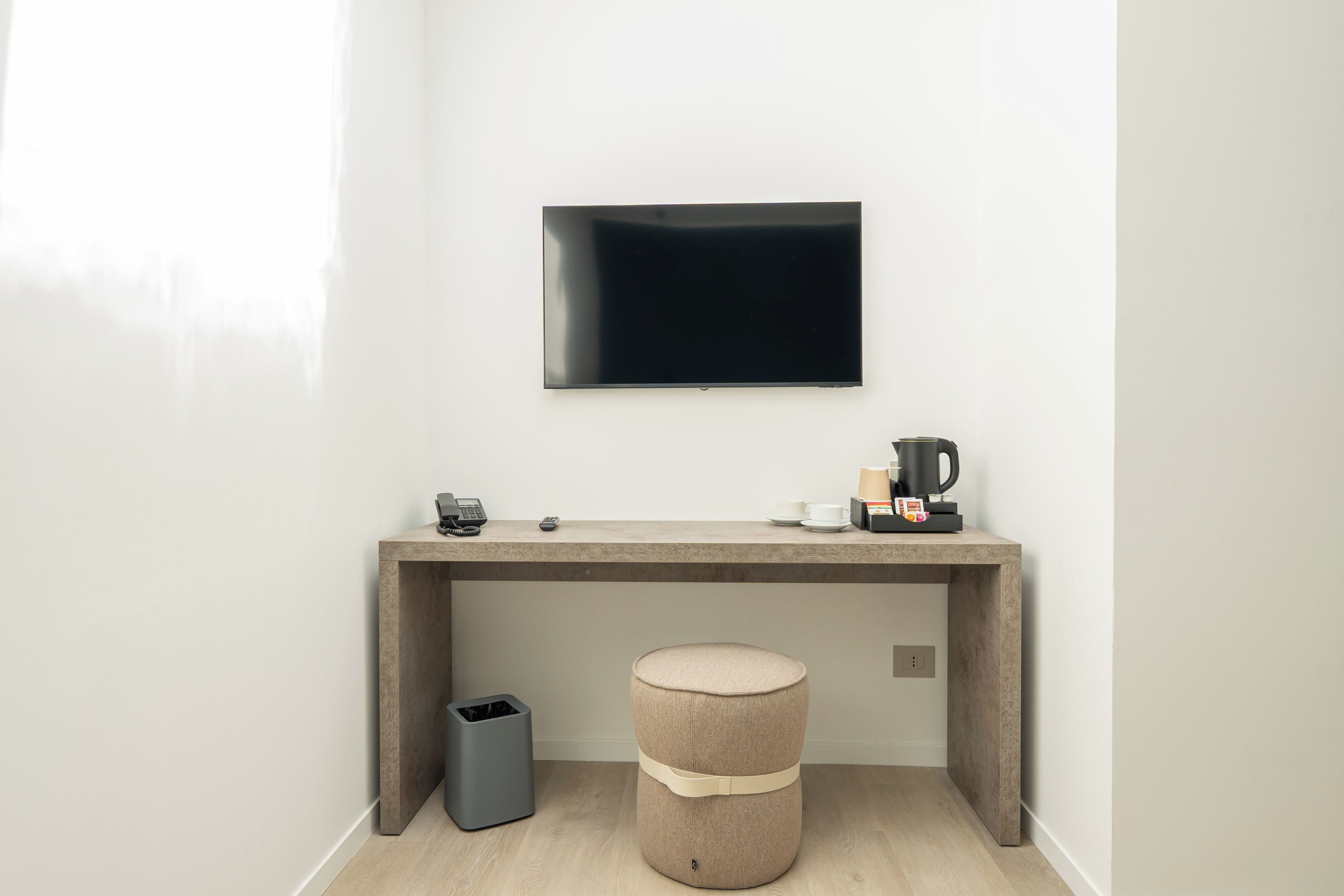 Une pièce au décor minimaliste, avec un téléviseur à écran plat fixé au mur, un petit bureau avec une machine à café, une poubelle et un pouf rond.