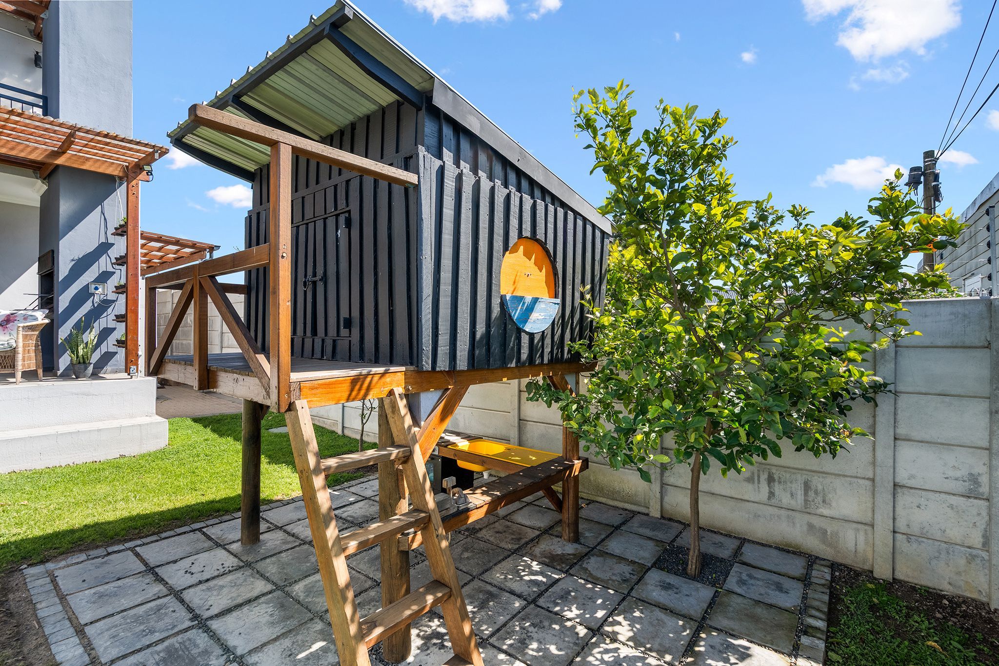 A wooden birdhouse with a sloped roof and a circular window, attached to a wooden structure with a ladder.