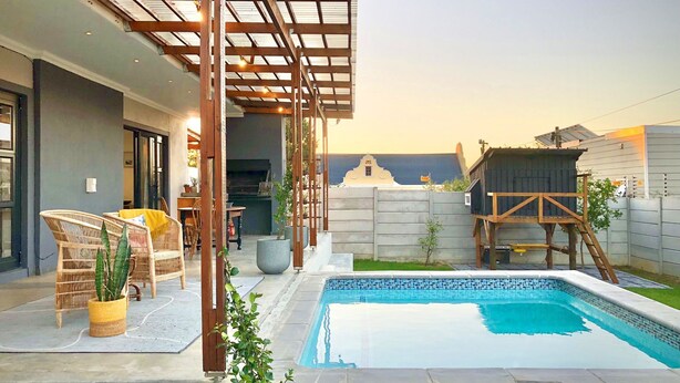 Een modern buitenzwembad met een pergola, rieten meubels en een klein speelhuisje.