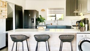 Een moderne keuken met een centraal eiland, barstoelen, een ingebouwde wasmachine en een raam met zonwering.
