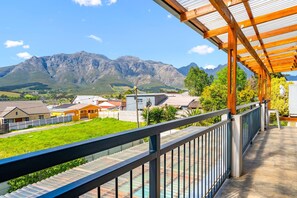 Un balcón con vistas a montañas, una pérgola de madera y una zona ajardinada con edificios al fondo.