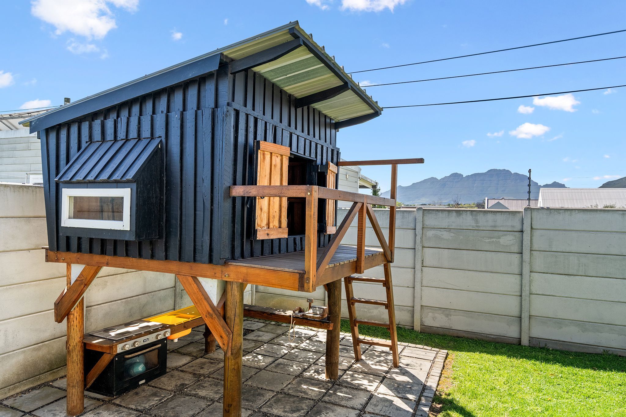 Una casita de madera con techo inclinado y una ventana, ubicada sobre una base de concreto con una escalera de madera.