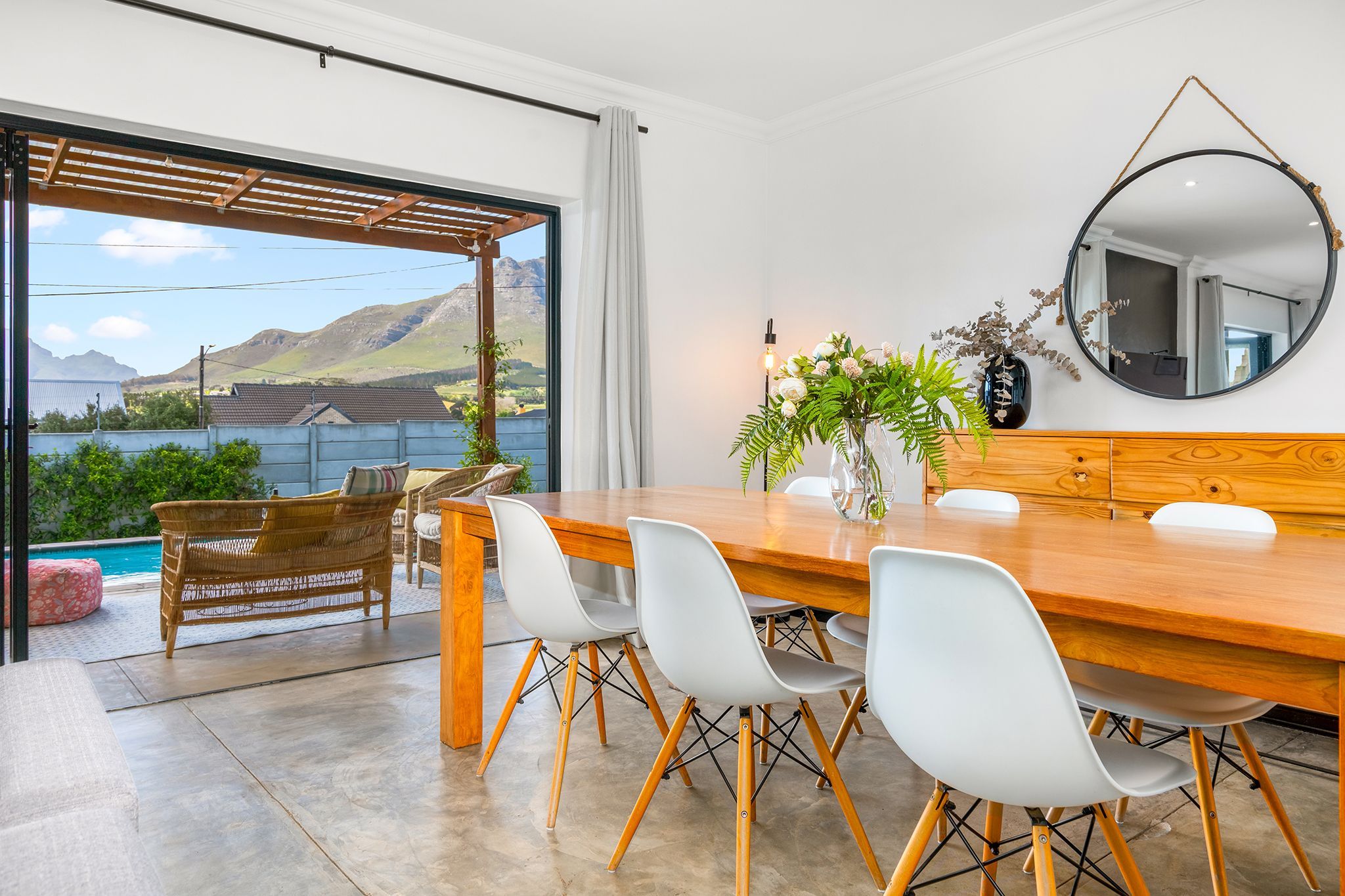 Un coin repas avec une table et des chaises en bois, un grand miroir et une vue sur les montagnes et une piscine.