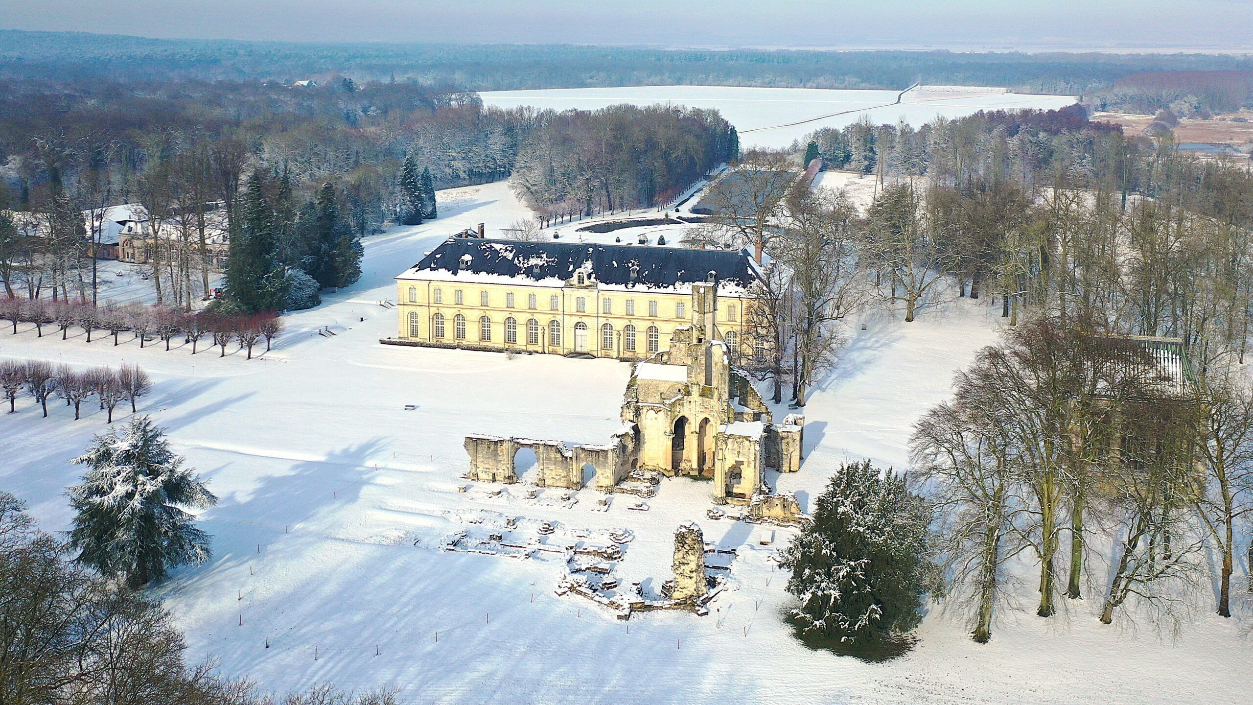 Een groot, historisch gebouw met een symmetrische gevel, omgeven door sneeuwbedekte velden en bomen.