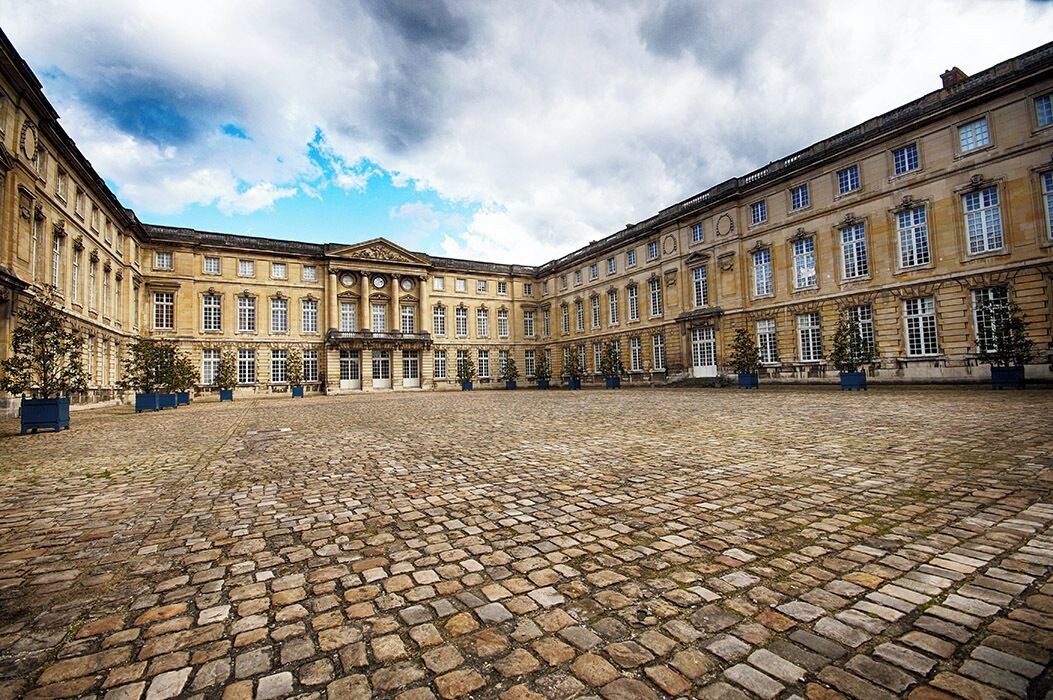 A courtyard with a cobblestone floor, large classical buildings with multiple windows, and potted plants.