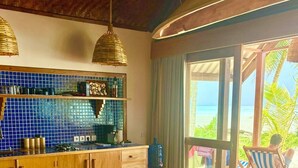 A room with a wooden bench, a kitchen area with blue tiles, and a view of the beach. - Ombak Biru Resort (Sipura Island)