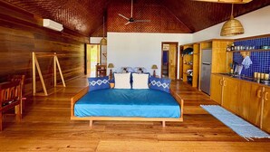 A spacious room with a large bed, wooden flooring, a ceiling fan, and a kitchen area with wooden cabinets. - Ombak Biru Resort (Sipura Island)