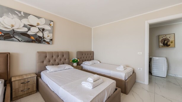 A hotel room with two beds, a nightstand, a small table with a vase of flowers, and a painting on the wall. - Sun Sea Apartment Durr s Waterfront (Durrës)