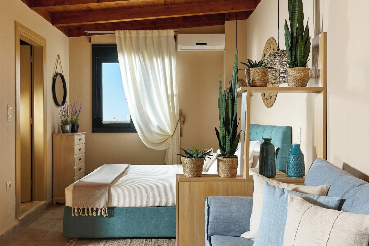 A bedroom with a bed, a wooden dresser, a window with curtains, a shelf with plants, and a blue sofa.
