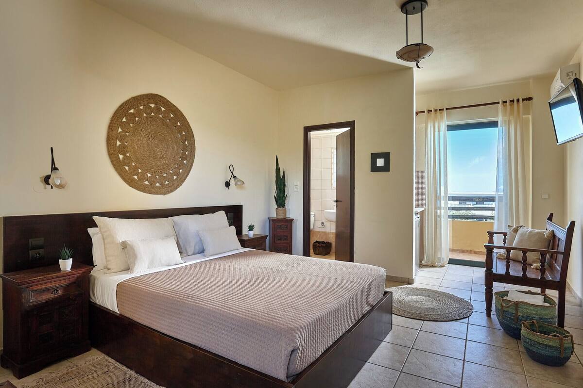 A bedroom with a bed, bedside tables, a wall-mounted lamp, a decorative wall piece, a potted plant, a chair, and a balcony with a view.