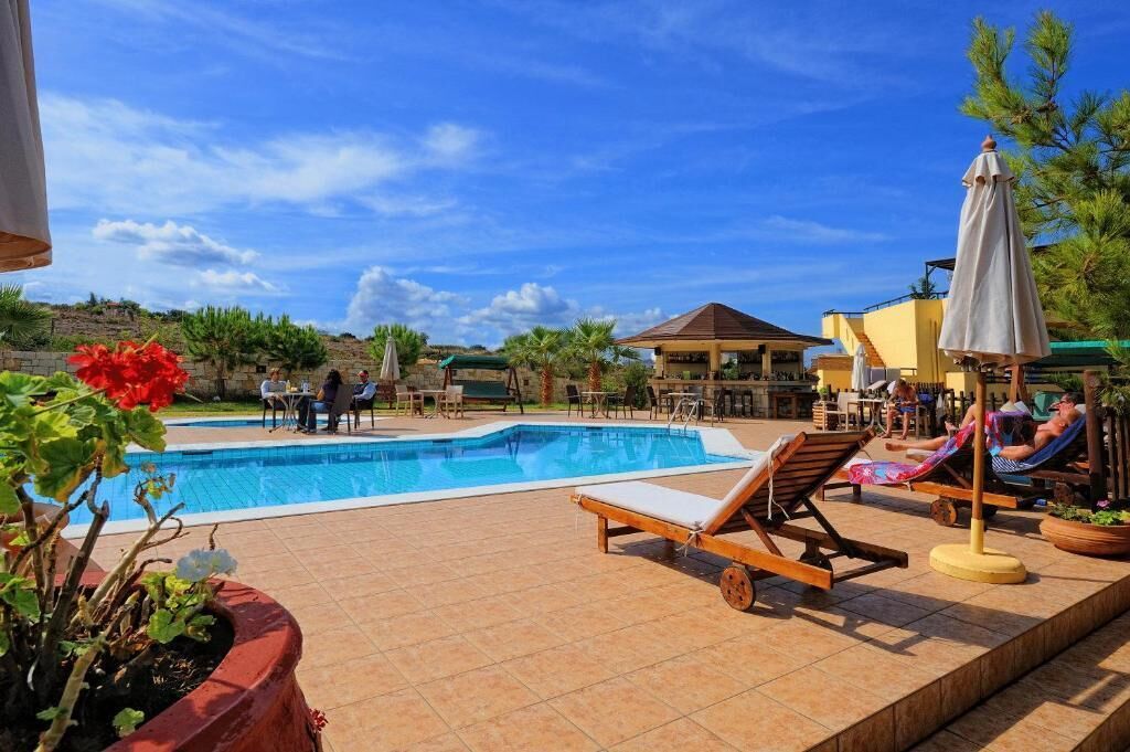 A pool area with lounge chairs and umbrellas, surrounded by a landscaped garden and a building with a roofed patio.