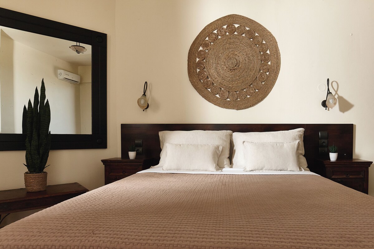 Una cama bien tendida con un edredón texturizado, un adorno circular tejido en la pared, un espejo grande y una planta en maceta sobre una mesita de noche.