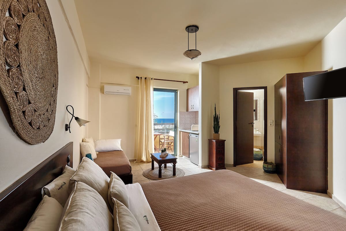 A hotel room with a bed, a desk, a chair, a large woven wall art piece, a ceiling light, and a view of the outside.