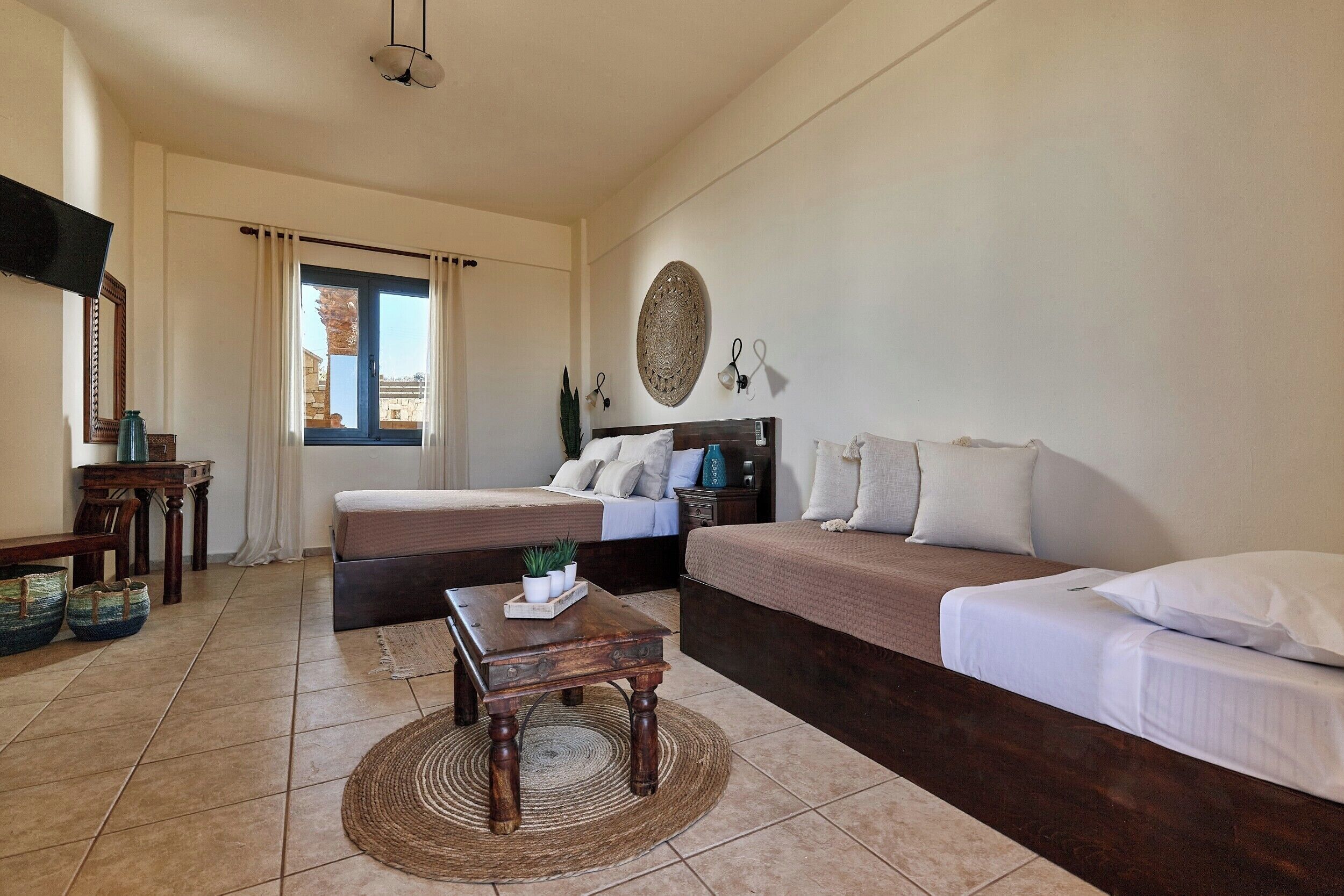 A bedroom with a bed, a sofa, a small table, a wall-mounted TV, and a window with curtains.