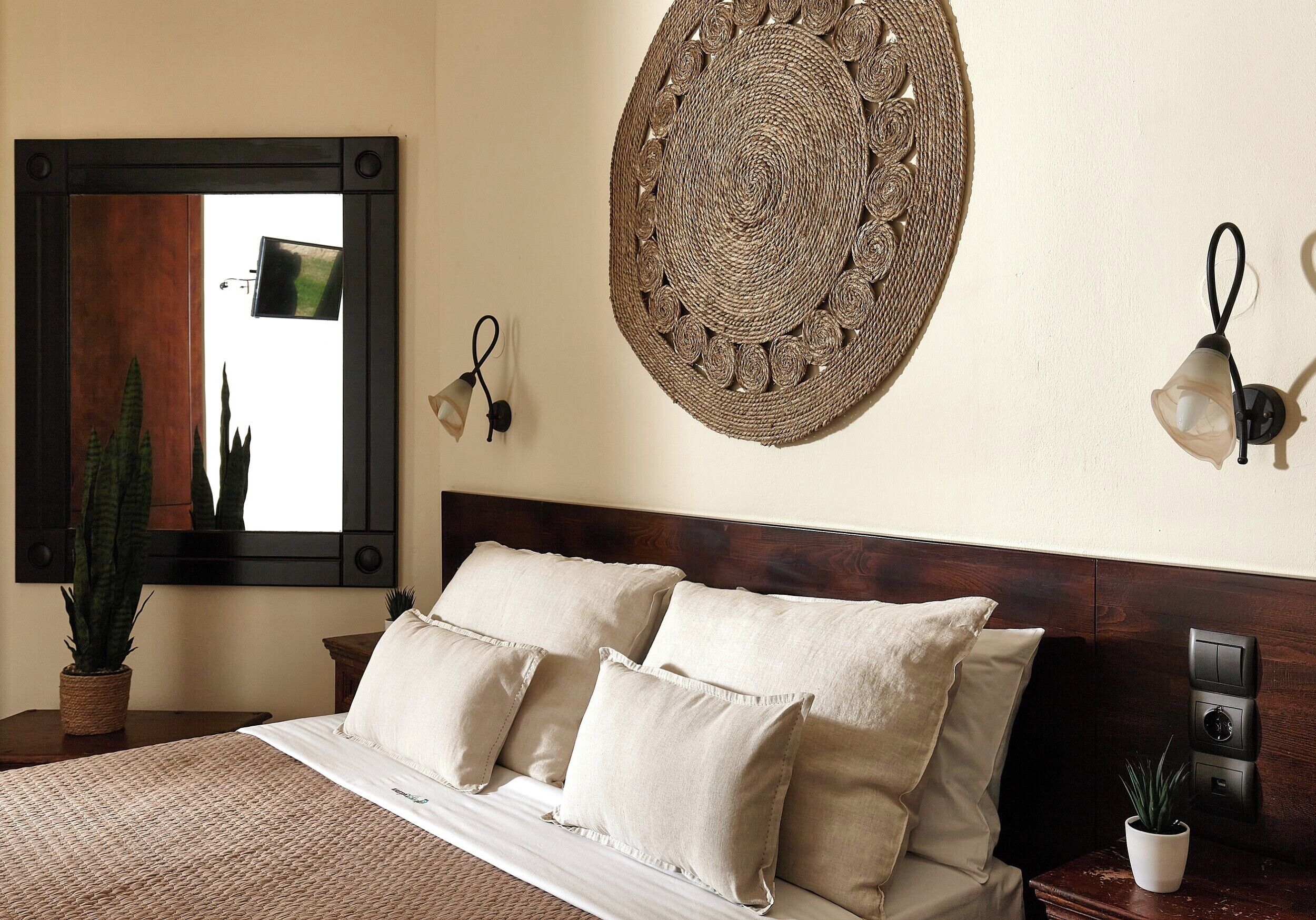 A neatly made bed with a headboard, a wall-mounted woven artwork, a potted plant, and a wall-mounted light fixture.