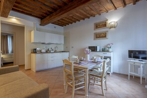 Een moderne keuken met witte kasten, een eettafel met stoelen, een televisie op een dressoir, en een plafond met zichtbare houten balken.