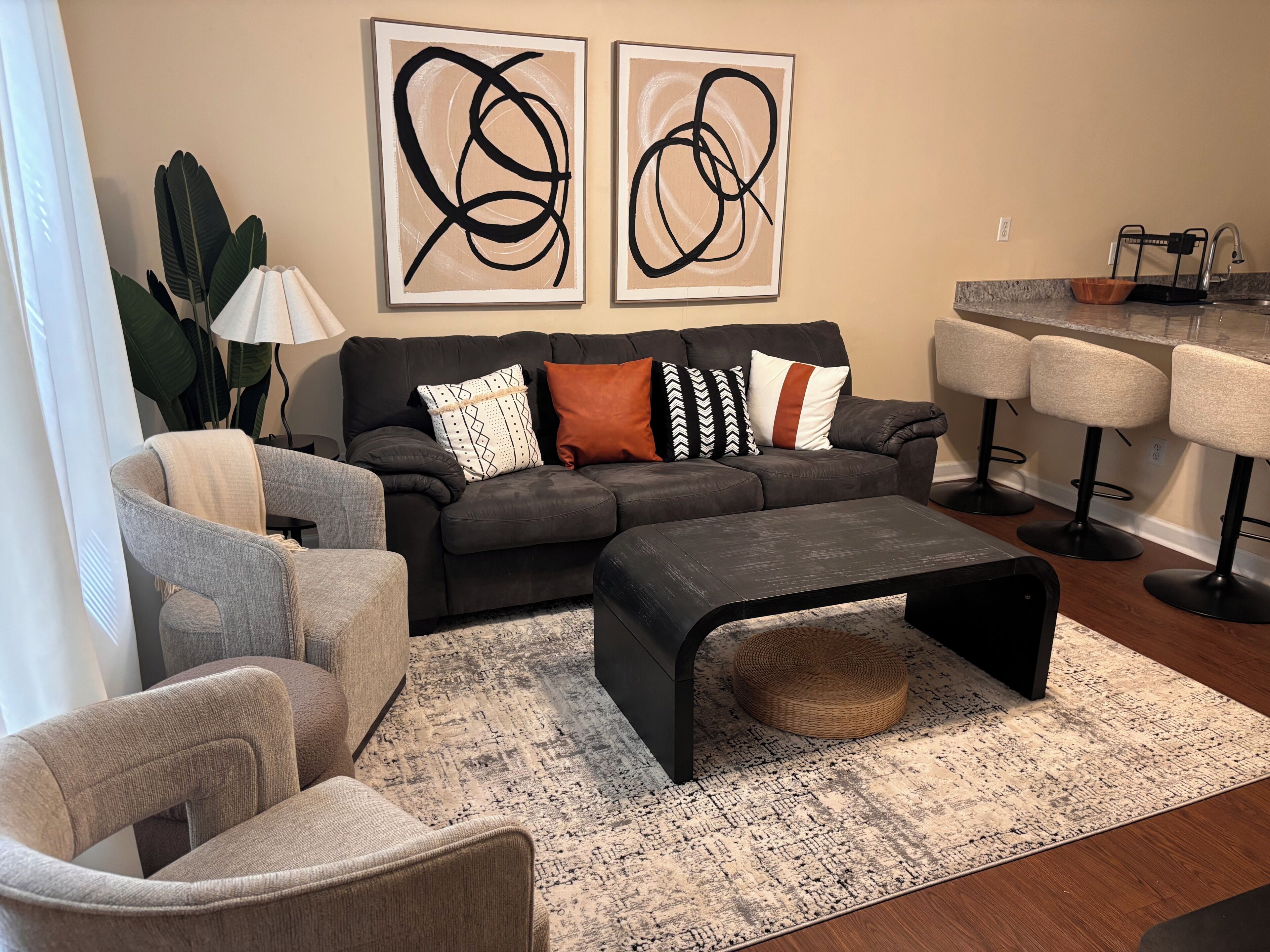 A living room with a sectional sofa, two armchairs, a coffee table, abstract wall art, a lamp, and a kitchen area with bar stools.