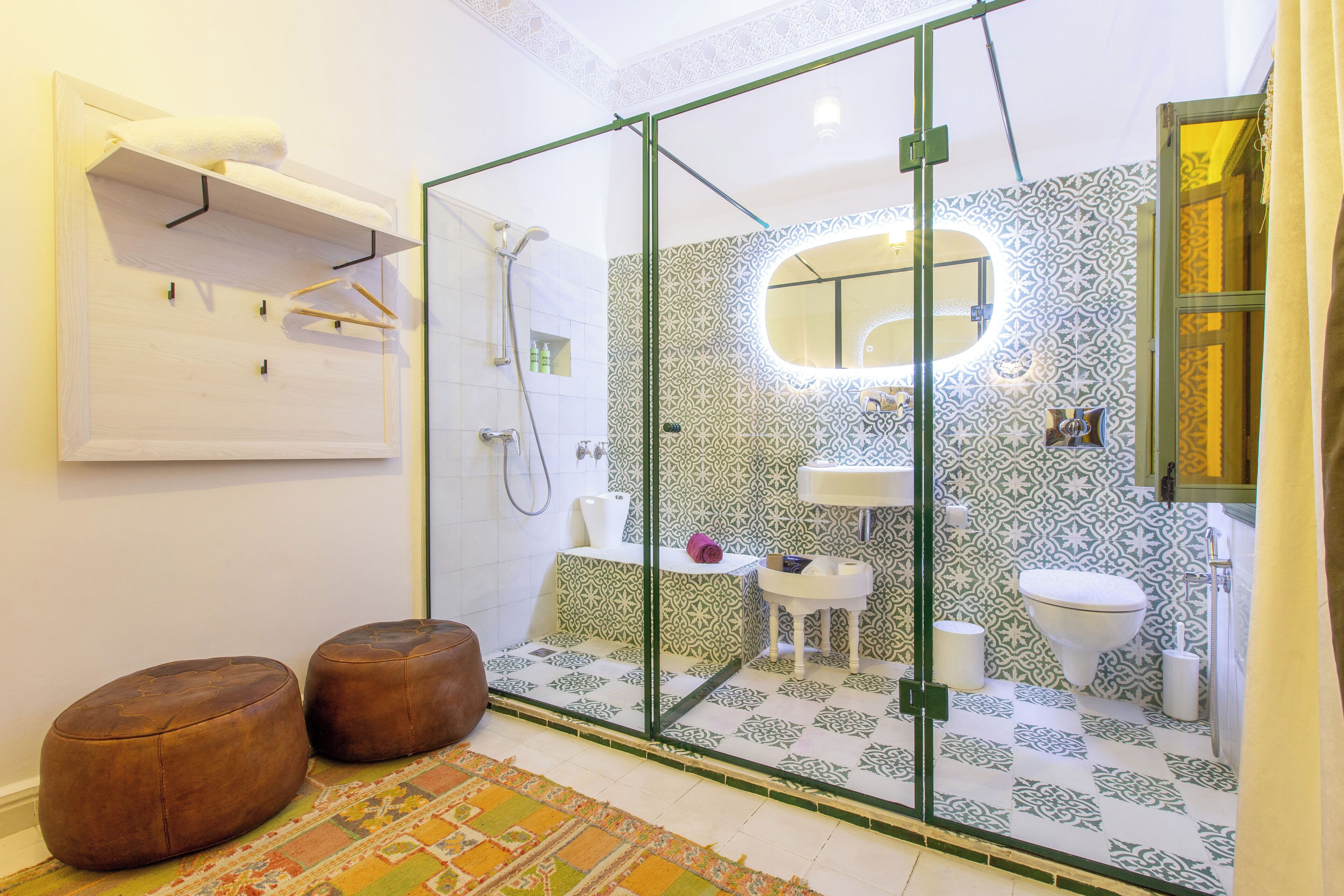 A bathroom with a glass shower enclosure, patterned tiles, a white sink, and two brown ottomans.