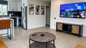 A modern kitchen with stainless steel appliances, a dining area with a round table, and a living room with a flat-screen TV displaying various channels. - Brand New next to Balboa Park, near Little Italy (San Diego)