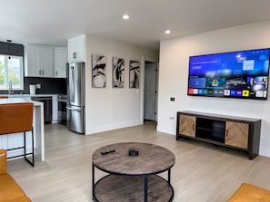 Una cocina moderna con electrodomésticos de acero inoxidable, un comedor con una mesa redonda y una sala de estar con un televisor de pantalla plana mostrando varios canales.