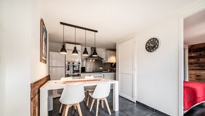 Eine moderne Küche mit Pendelleuchten, ein Essbereich mit weißen Stühlen, eine Uhr an der Wand und ein rotes Bett im angrenzenden Zimmer.