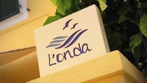 Une enseigne portant le nom « L’onda », sur un fond blanc, est fixée à une structure jaune, avec un feuillage vert en arrière-plan.