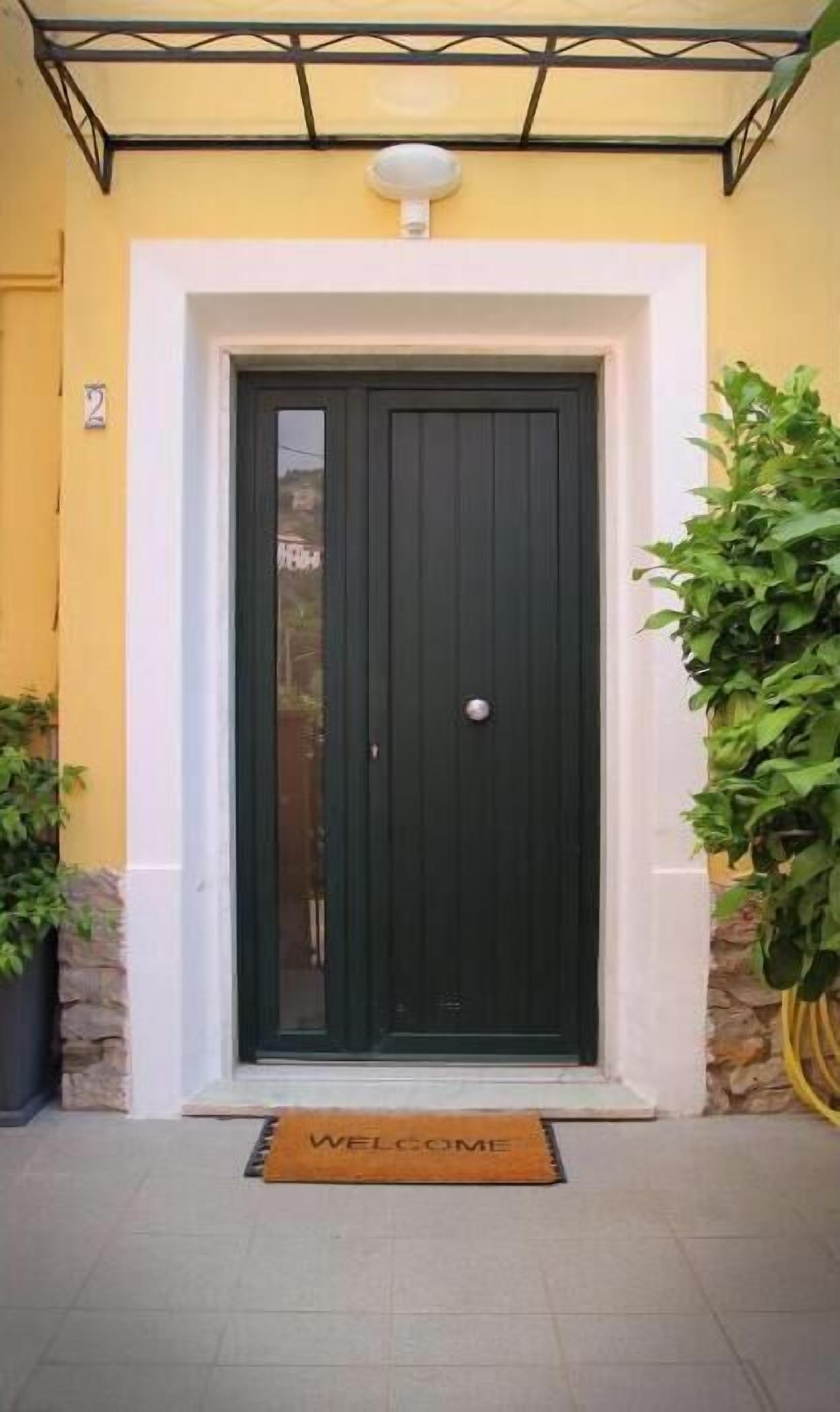 A black door with a white doorbell, a welcome mat, and potted plants on either side.