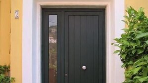 Eine schwarze Tür mit weißem Klingelknopf, einer Fußmatte und je einem Blumentopf an jeder Seite.