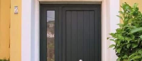 A black door with a white doorbell, a welcome mat, and potted plants on either side.