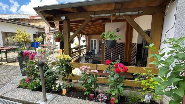 A covered outdoor seating area with wooden benches, potted plants, and a view of a building in the background. - Pezion and apartman near Karlovy Vary (Chodov)