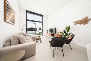 Ein modernes Wohnzimmer mit einer Couch, zwei Sesseln, einem Couchtisch, einem Fernseher und einem großen Fenster.