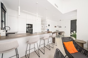 Eine moderne Küche mit weißen Schränken, einer zentralen Kochinsel, Barhocker, einem Essbereich mit Tisch und Stühlen sowie einem Korbstuhl mit orangefarbenem Kissen.