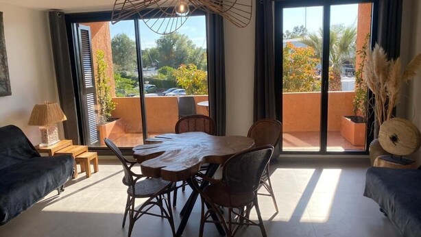 A room with a dining table, chairs, a sofa, a lamp, and a view through large windows. - Mer et soleil appartements (Cogolin)