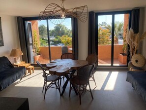A room with a dining table, chairs, a sofa, a lamp, and a view through large windows. - Mer et soleil appartements (Cogolin)