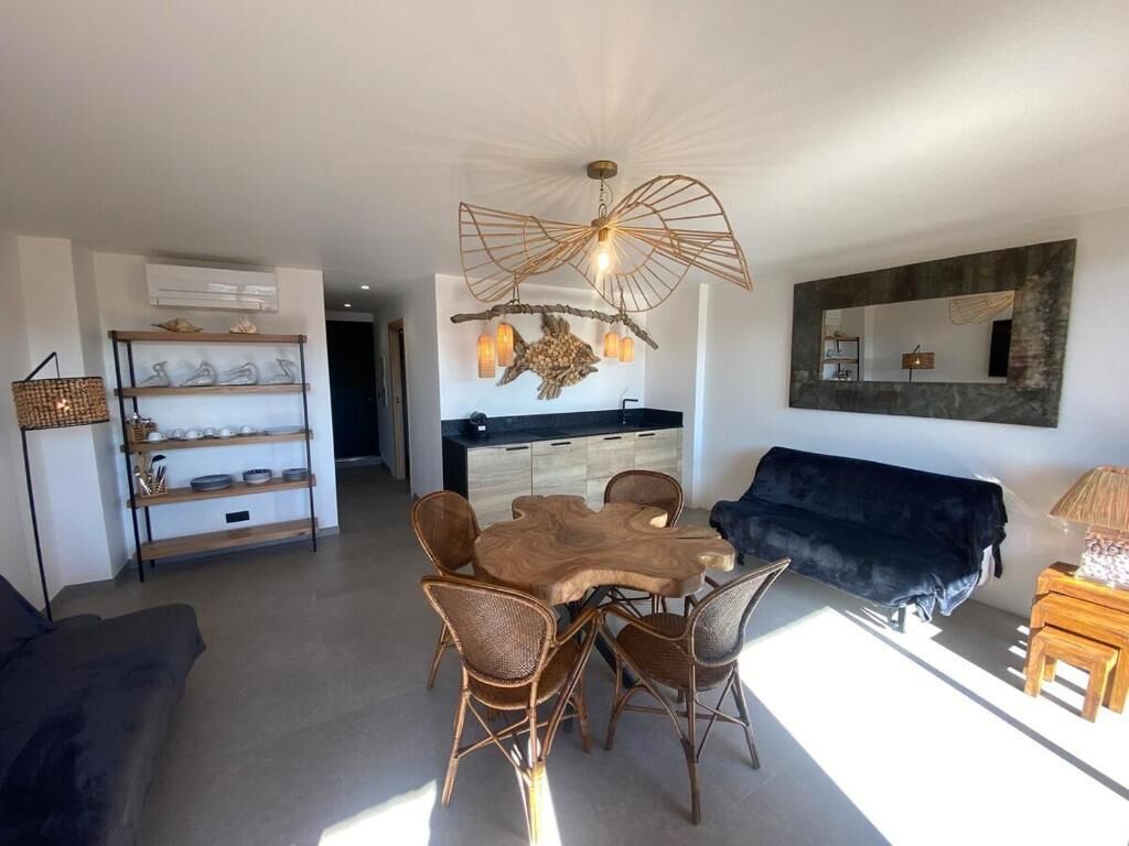 A modern living room with a dining table, wicker chairs, a dark sofa, a unique ceiling light, and a kitchen in the background.