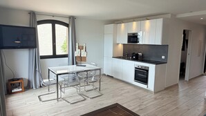 A modern kitchen with white cabinets, a microwave, and an oven. There is a dining table with chairs and a TV mounted on the wall. - Mer et soleil appartements (Cogolin)